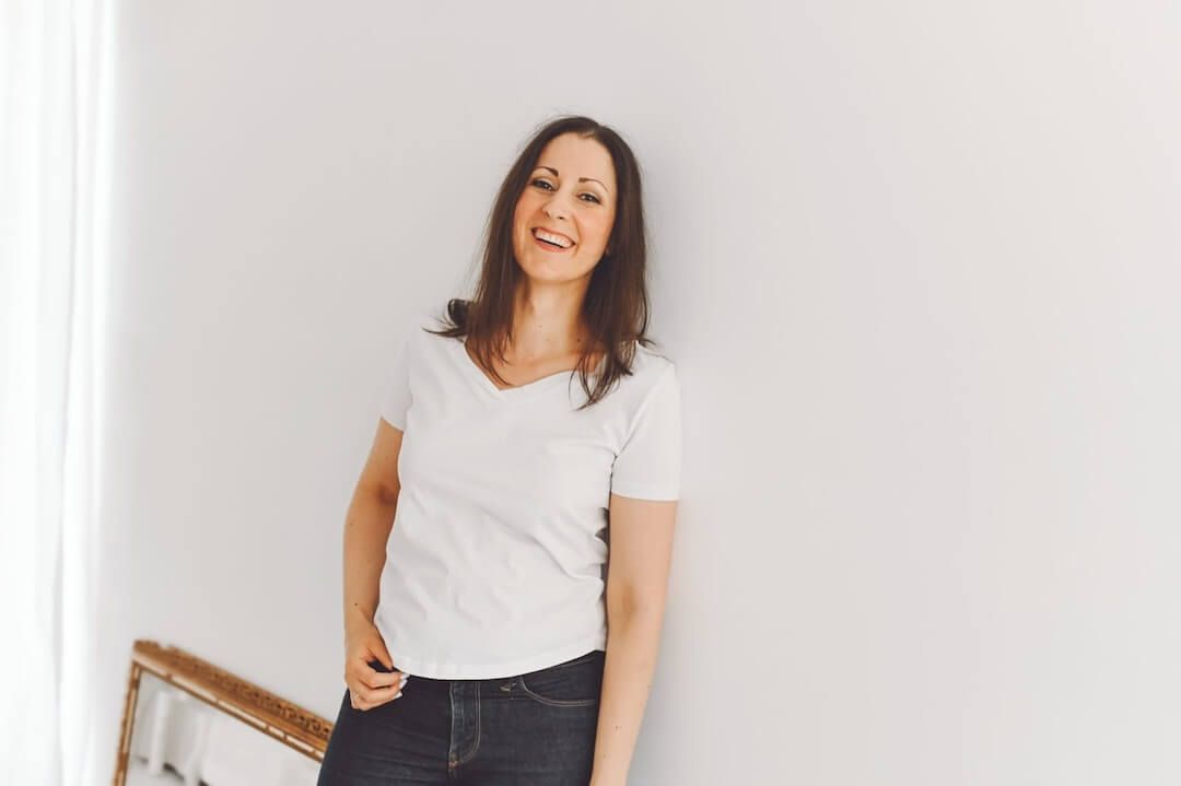 coaching Una mujer sonriente con el pelo largo y oscuro se alza contra una pared blanca. Lleva una camiseta blanca informal con cuello en V y jeans oscuros. La esquina de un marco de madera es visible en la parte inferior izquierda de la imagen.