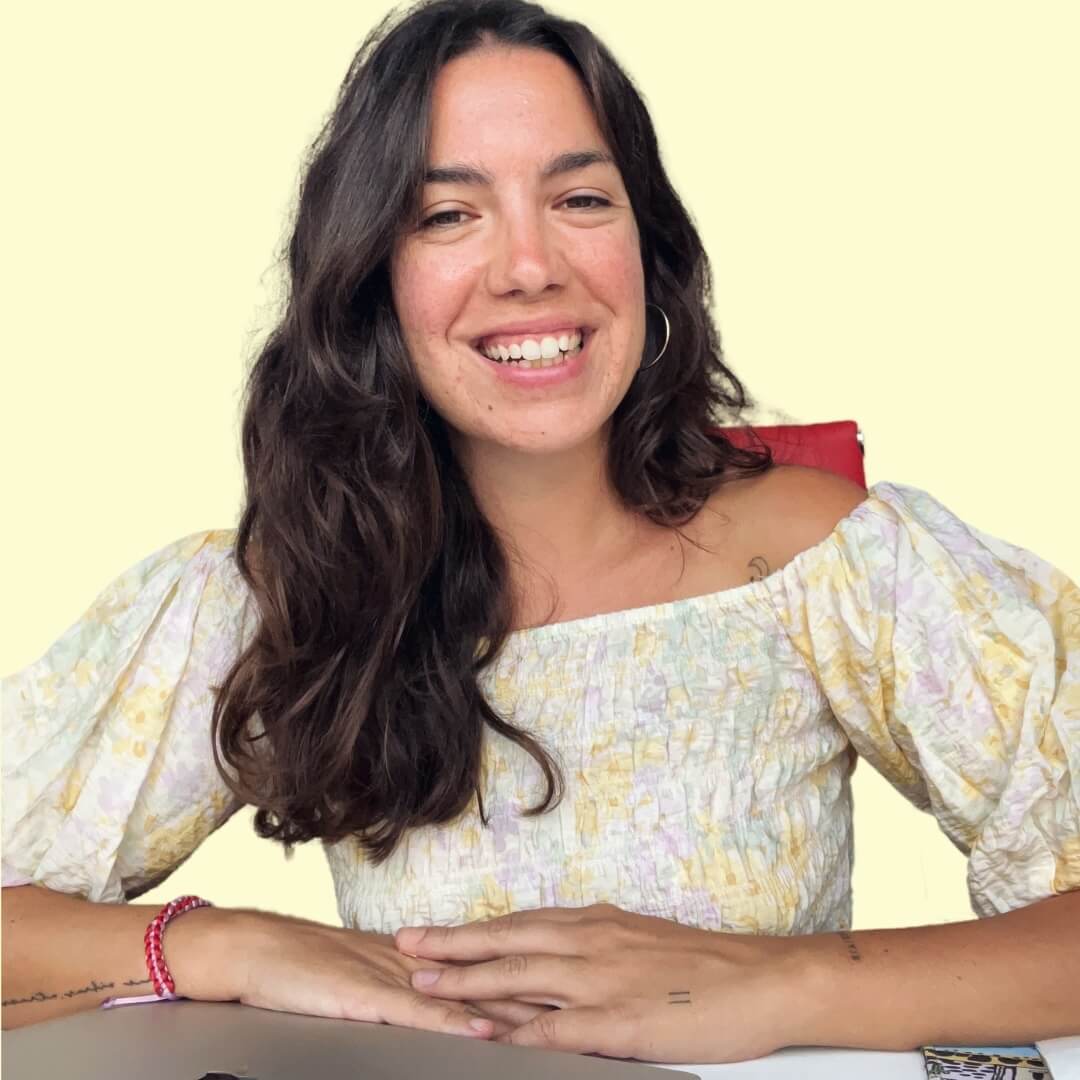 coaching Una mujer con cabello castaño largo y ondulado sonríe a la cámara. Lleva una blusa de color claro con hombros descubiertos y mangas abullonadas. Sus manos descansan sobre una superficie plana. Tiene una expresión relajada y alegre, con un fondo liso de color amarillo claro detrás de ella. spiritual mood