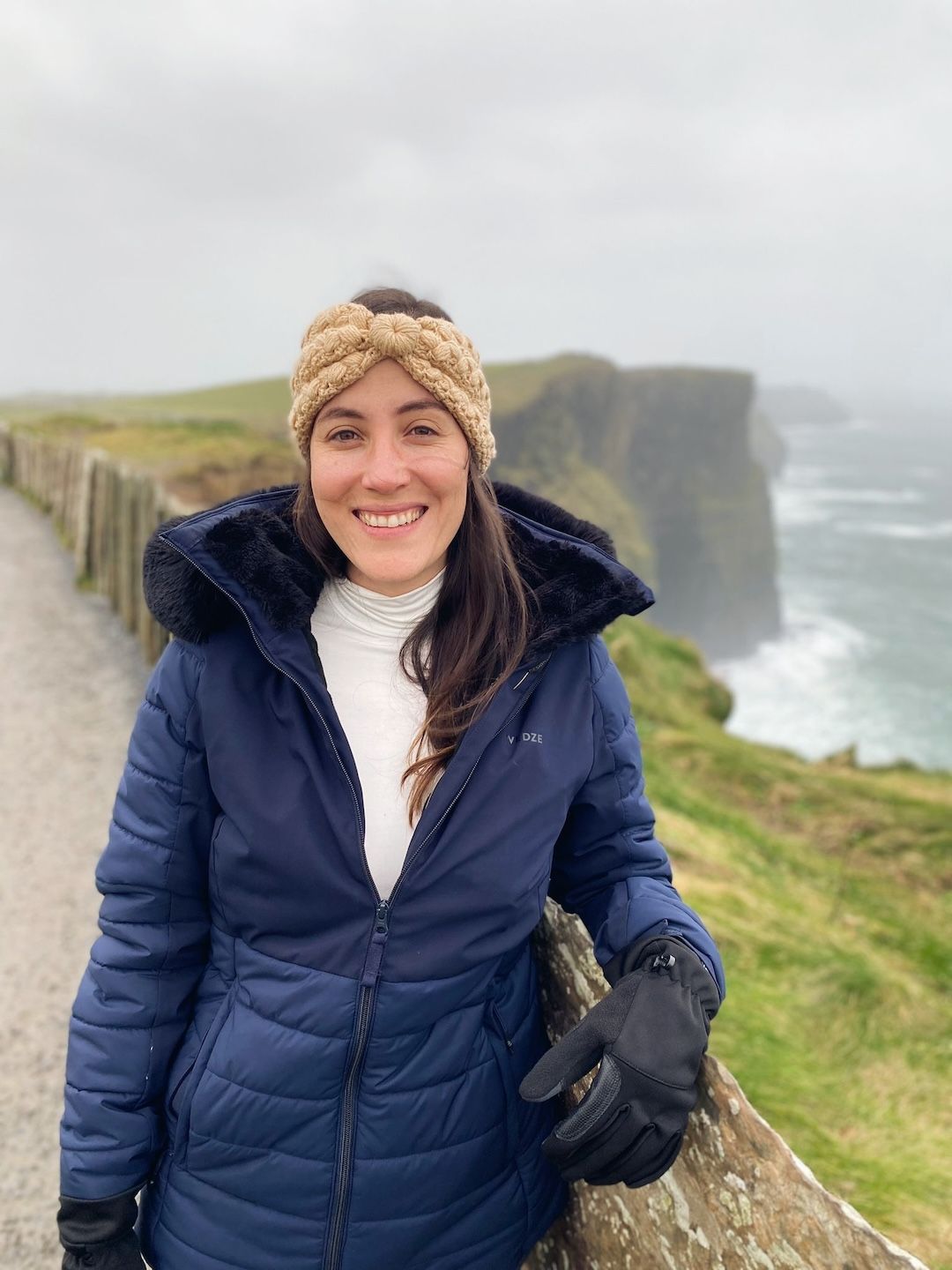 coaching Una mujer sonríe junto a una valla de madera con los acantilados de Moher al fondo en un día nublado. Lleva una chaqueta azul, guantes y una diadema tejida. Las olas del océano chocan contra los acantilados detrás de ella.