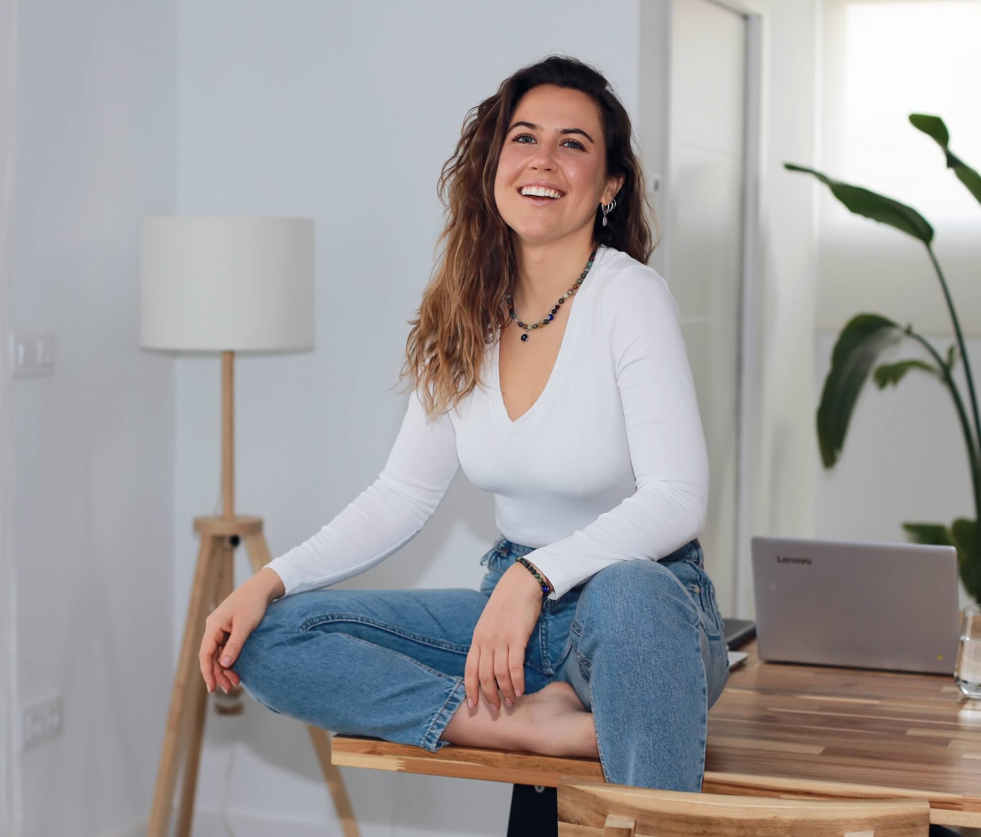 Alejandra Rodriguez. coaching Una mujer de cabello ondulado, vestida con una camisa blanca de manga larga y jeans azules, está sentada con las piernas cruzadas sobre una mesa de madera, sonriendo. Al fondo, hay una lámpara de pie, un ordenador portátil sobre la mesa, una planta y una pared blanca. spiritual mood