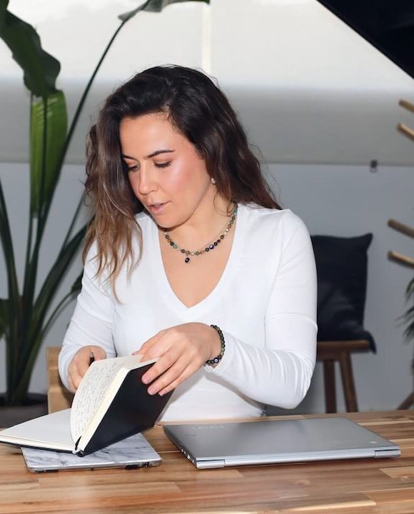 Alejandra Rodríguez. coaching Una persona con el pelo largo y una blusa blanca está sentada en una mesa de madera hojeando un cuaderno. Sobre la mesa hay un portátil cerrado y un vaso de agua. El fondo incluye plantas en macetas y una escalera. spiritual mood
